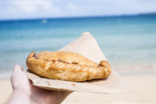A Cornish pasty on a paper bag held out by a hand with the sea and beach in the background.