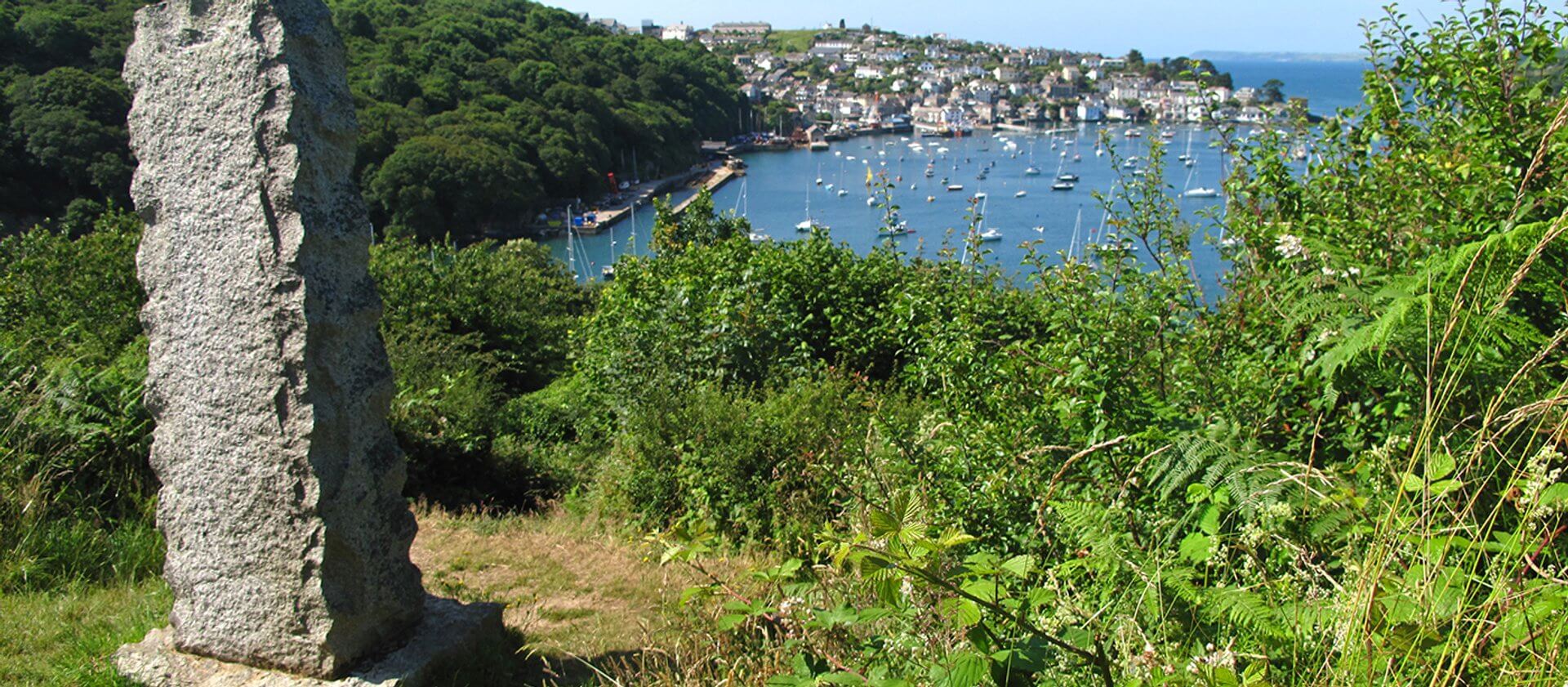 The view of Fowey from the Fowey Hall Walk, with greenery in the forground and a pillar rock with the estuary below full of boats and the houses on the opposite bank.