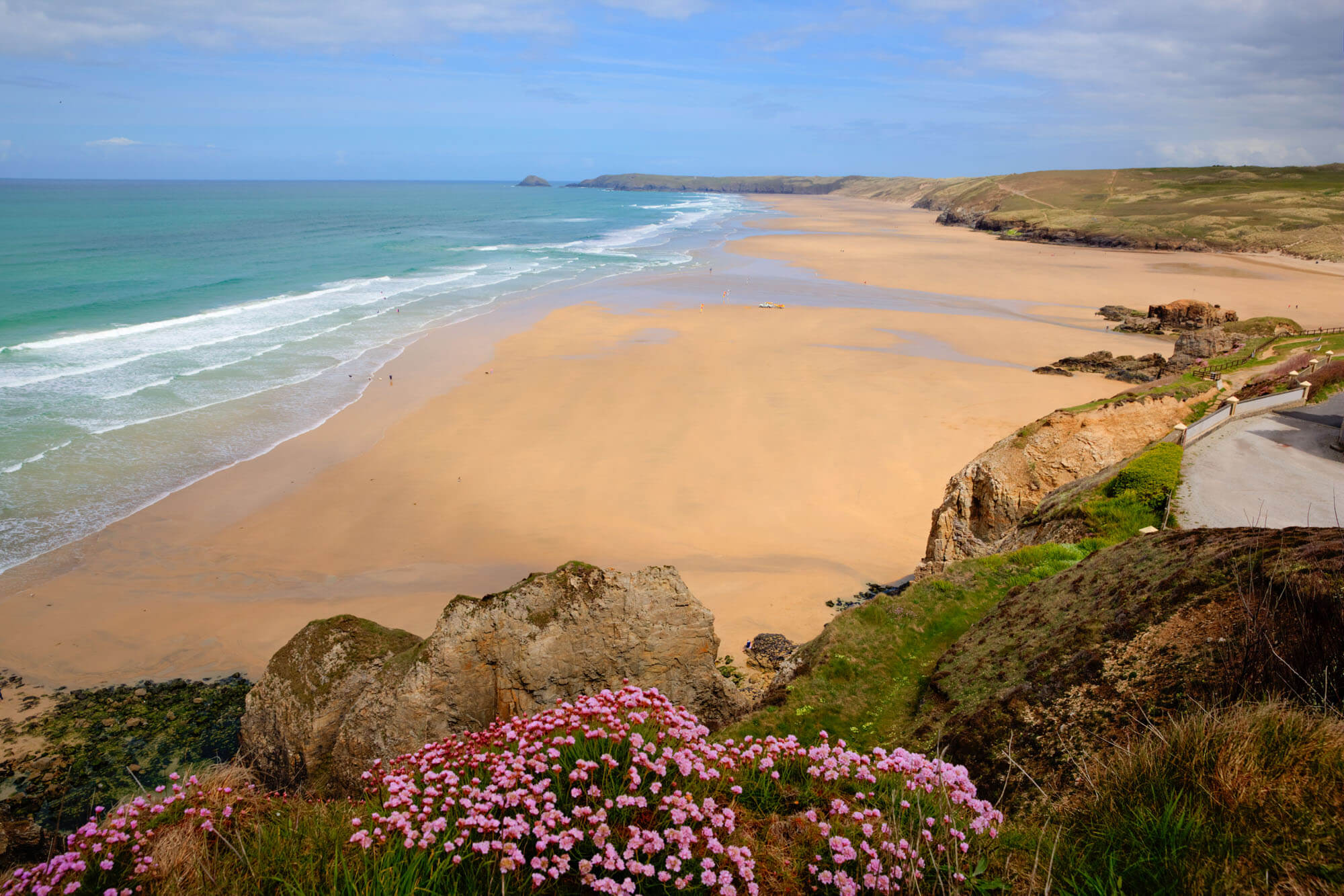 The view from the coastal path across Perranporth beach, with a huge sandy shore and flowers on the rocky cliffs.